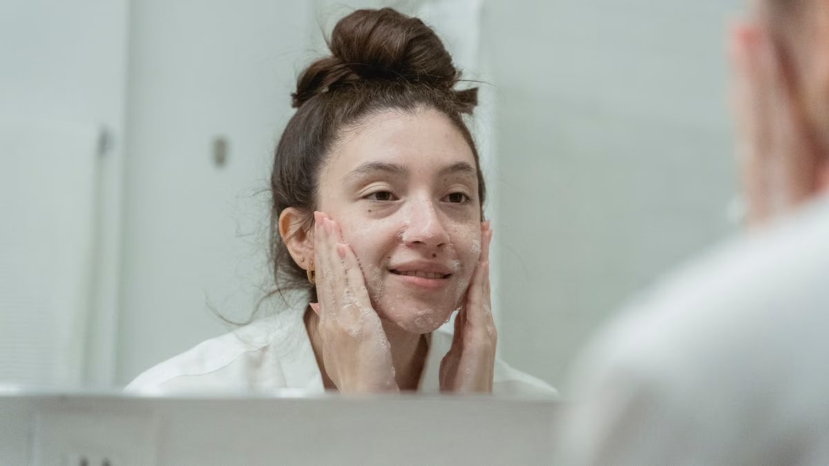 A woman smiling as she applies facial cream in front of a bathroom mirror, enhancing skincare routine.